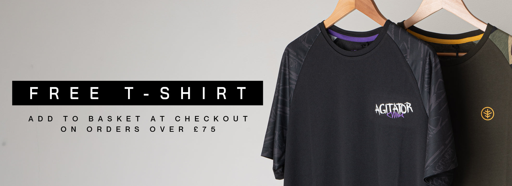 Two short-sleeve t-shirts hanging on wooden hangers against a light gray background. The front shirt is black with a purple collar lining and a white 'AGITATOR' logo on the chest. Theback top is green with Wychwood Logo